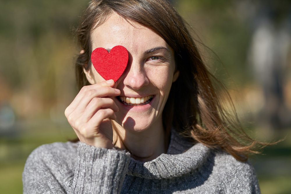 Eine junge Frau hält sich ein rotes Filzherz vor ihr Auge und lächelt.